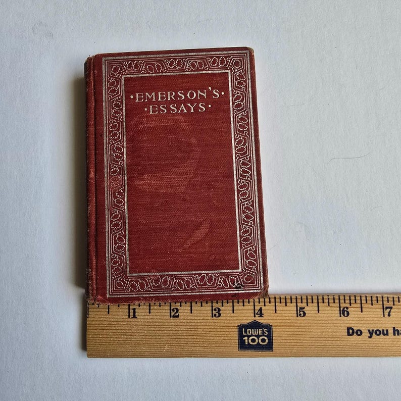 Peut inclure: Un petit livre ancien intitul&eacute; "Emerson's Essays" avec une couverture rouge et une bordure argent&eacute;e. Le livre est plac&eacute; &agrave; c&ocirc;t&eacute; d'une r&egrave;gle en bois, indiquant sa taille. La couverture du livre est l&eacute;g&egrave;rement us&eacute;e, ce qui sugg&egrave;re son &acirc;ge.