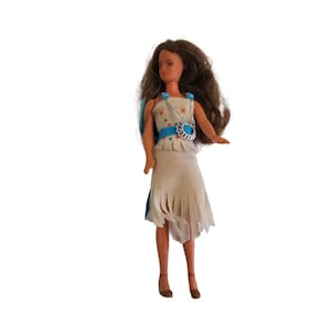 May include: A doll with long brown hair wearing a white dress with blue trim and a turquoise pendant necklace.
