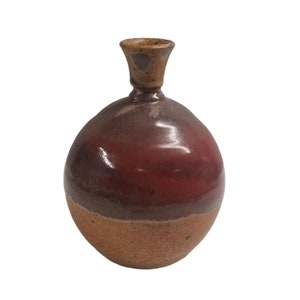 Könnte beinhalten: Eine handgefertigte Keramikvase mit rundem Körper und schmalem Hals. Die Vase zeigt einen Farbverlauf, der von einem hellbraunen Sockel zu einem tiefroten Mittelteil und einem dunkleren braunen Hals übergeht. Die Vase hat eine glänzende Oberfläche.