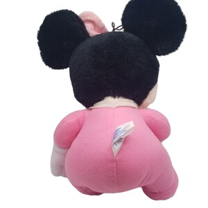 Puede incluir: Un juguete de peluche rosa Minnie Mouse con orejas negras y un lazo blanco. El juguete lleva un atuendo rosa con detalles blancos.