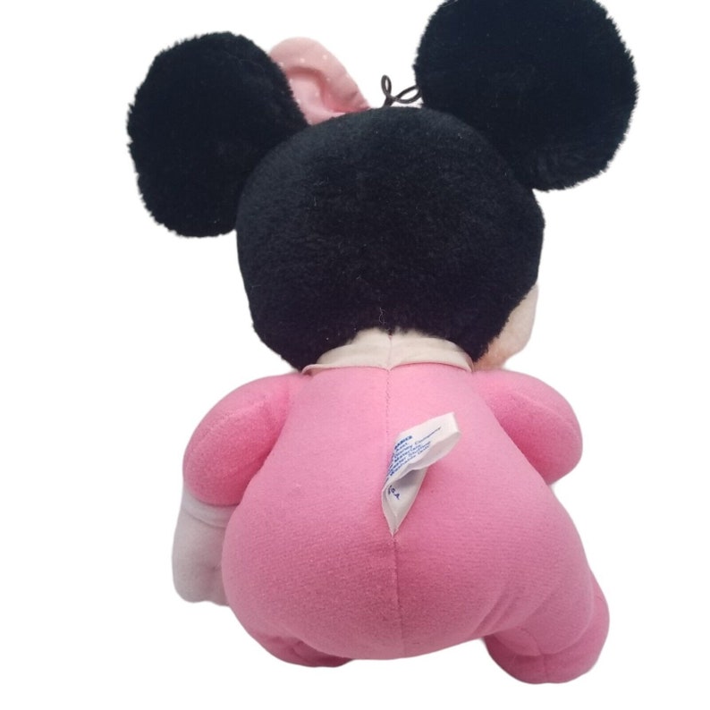 Puede incluir: Un juguete de peluche rosa Minnie Mouse con orejas negras y un lazo blanco. El juguete lleva un atuendo rosa con detalles blancos.