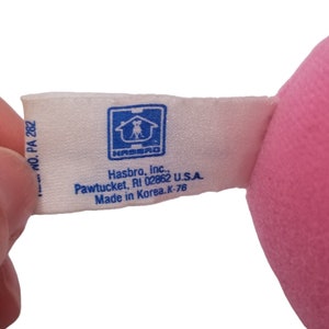 Puede incluir: Una etiqueta blanca con texto azul y un logotipo azul de Hasbro. La etiqueta dice "Hasbro, Inc., Pawtucket, RI 02862 U.S.A. Hecho en Corea. K-76"