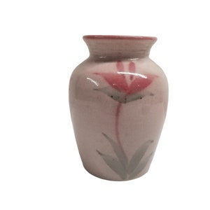 Puede incluir: Un jarrón pequeño de cerámica rosa con un diseño floral. El jarrón tiene una sola flor rosa con hojas verdes pintadas en la parte delantera.