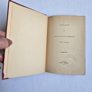 Peut inclure: Livre ouvert avec une couverture rouge, affichant le titre "ESSAYS" de Ralph Waldo Emerson. Le texte "FIRST SERIES" est en dessous. Un tampon de "PHILADELPHIA LIBRARIES" est visible.