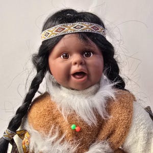 May include: A Native American doll with dark hair in braids and a beaded headband. The doll has brown eyes and is wearing a brown and white outfit with a fur-like collar. The doll's mouth is open, and it appears to be smiling.