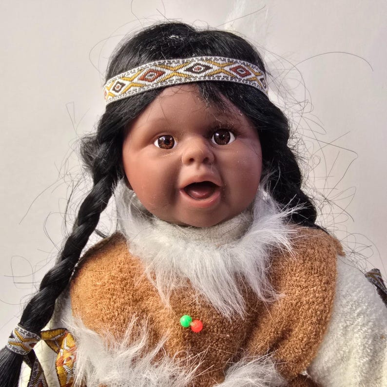May include: A Native American doll with dark hair in braids and a beaded headband. The doll has brown eyes and is wearing a brown and white outfit with a fur-like collar. The doll's mouth is open, and it appears to be smiling.