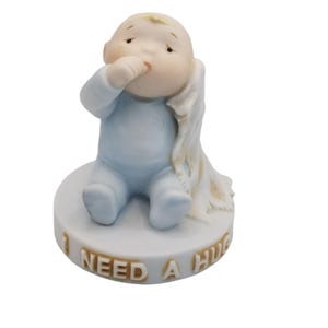May include: A ceramic figurine of a baby in a light blue onesie, sitting on a white base. The baby is sucking its thumb and has a blanket draped over its shoulder. The base reads "I NEED A HUG".
