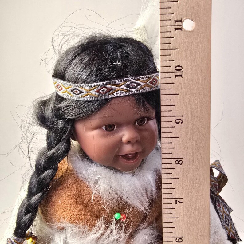 May include: A Native American-style doll with dark hair in a braid, wearing a brown and white outfit with a beaded headband. The doll has a smiling expression. A wooden ruler is next to the doll.