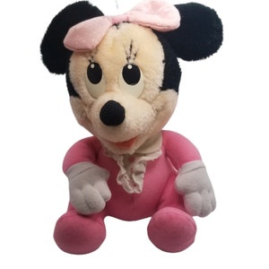 Puede incluir: Un juguete de peluche rosa Minnie Mouse con un lazo rosa con lunares blancos. El juguete tiene orejas negras y una nariz negra. El juguete est&aacute; sentado con las piernas cruzadas y los brazos extendidos.