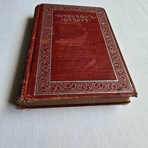 Peut inclure: Un livre ancien avec une couverture rouge et des lettres et bordures argent&eacute;es. Le titre "EMERSON'S ESSAYS" est visible sur la couverture. Le livre montre des signes d'usure, sugg&eacute;rant son &acirc;ge et son utilisation. Le fond est une surface blanche unie.