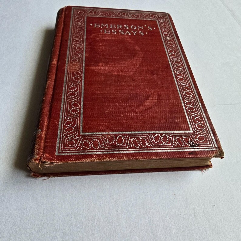 Peut inclure: Un livre ancien avec une couverture rouge et des lettres et bordures argent&eacute;es. Le titre "EMERSON'S ESSAYS" est visible sur la couverture. Le livre montre des signes d'usure, sugg&eacute;rant son &acirc;ge et son utilisation. Le fond est une surface blanche unie.