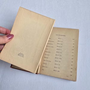 Peut inclure: Un livre ancien ouvert, avec une tranche rouge et des pages cr&egrave;me vieilles. La page de gauche montre un tampon circulaire en relief avec du texte, tandis que la page de droite affiche une table des mati&egrave;res avec des titres et des num&eacute;ros de page. La couverture du livre est l&eacute;g&egrave;rement soulev&eacute;e.