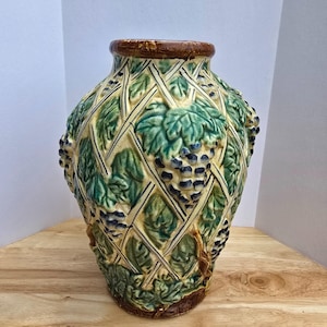 May include: A decorative ceramic vase with a textured surface. The vase features a lattice design with green leaves and clusters of blue grapes. The rim and base are brown.