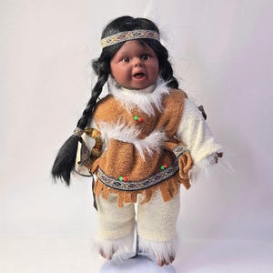 May include: A Native American doll with dark skin, black braided hair, and a patterned headband. The doll wears a brown vest with white faux fur trim, a white long-sleeved shirt, and off-white pants with faux fur trim at the ankles. The doll is standing.