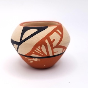 Puede incluir: Una pequeña olla de cerámica pintada a mano con un diseño geométrico en negro, blanco y naranja. La olla tiene una forma redondeada y un borde ligeramente acampanado.