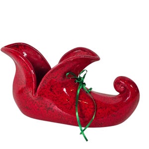 Puede incluir: Un zapato de elfo de cerámica roja brillante con un lazo de cinta verde. El zapato tiene una textura moteada y una punta rizada. La parte superior abierta sugiere que podría usarse como un recipiente decorativo o una jardinera.