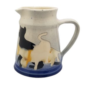 Puede incluir: Una jarra de cerámica con un diseño de tres gatos azules, blancos y negros. La jarra tiene un asa y un cuerpo redondeado.