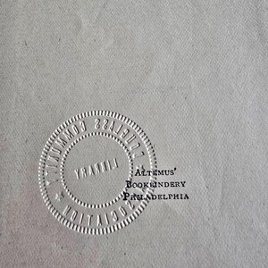 Peut inclure: Gros plan d'une surface gris clair avec un motif circulaire en relief. Le motif comprend le texte "COMMUNITY LIBRARY" et "1902" sur le pourtour. Le texte "ALTEMUS' BOOKBINDERY PHILADELPHIA" est &eacute;galement visible.