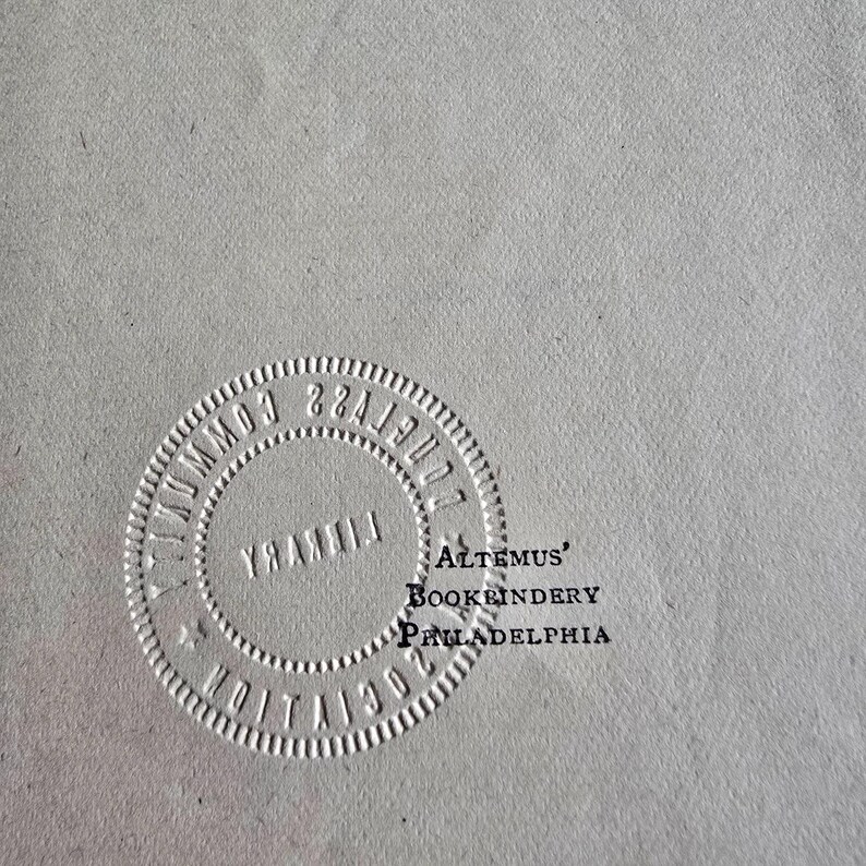 Peut inclure: Gros plan d'une surface gris clair avec un motif circulaire en relief. Le motif comprend le texte "COMMUNITY LIBRARY" et "1902" sur le pourtour. Le texte "ALTEMUS' BOOKBINDERY PHILADELPHIA" est &eacute;galement visible.