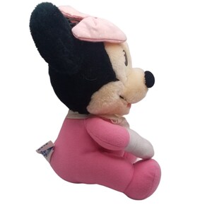 Puede incluir: Un juguete de peluche rosa Minnie Mouse con un sombrero rosa con lunares blancos y un atuendo rosa. El juguete est&aacute; sentado con las piernas cruzadas y tiene un lazo blanco en la cabeza.