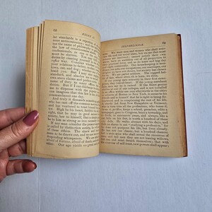 Peut inclure: Livre ouvert avec des pages jaunies et vieillies. La page de gauche affiche le num&eacute;ro 68 et le titre "ESSAY II". La page de droite affiche le num&eacute;ro 69 et le titre "SELF-RELIANCE". Le livre est tenu ouvert par une main.