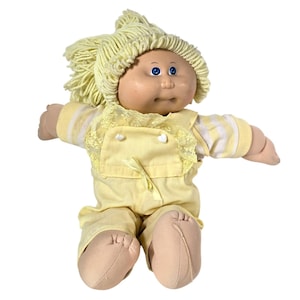 May include: A vintage Cabbage Patch Kid doll with a yellow yarn hairstyle and blue eyes. The doll wears a yellow outfit with white stripes and lace trim. The doll's skin is a light peach color.