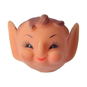 Puede incluir: Una cabeza de muñeca de goma vintage con una expresión sonriente. La cabeza de la muñeca tiene orejas grandes, una nariz larga y rasgos pintados, incluyendo pestañas, cejas y una boca roja. El pelo es castaño y está peinado en un pequeño remolino.