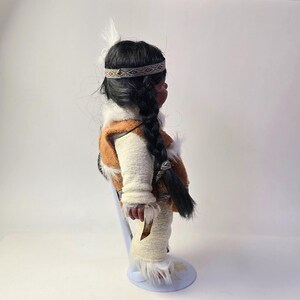 May include: A Native American doll with dark hair in a long braid, wearing a brown and white vest, cream-colored pants, and a patterned headband. The doll has a feather in its hair and is standing on a light blue stand.