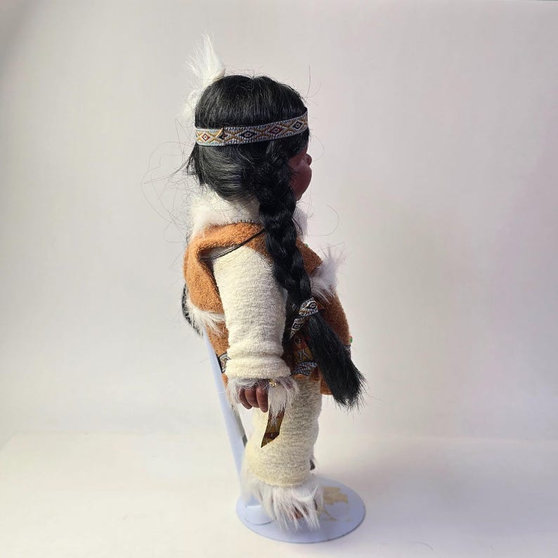 May include: A Native American doll with dark hair in a long braid, wearing a brown and white vest, cream-colored pants, and a patterned headband. The doll has a feather in its hair and is standing on a light blue stand.