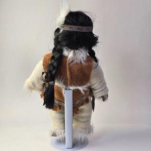 May include: A Native American doll, viewed from the back, with long black braided hair and a feather. The doll wears a cream-colored outfit with brown accents, a patterned headband, and a decorative belt. The doll is standing on a white stand.