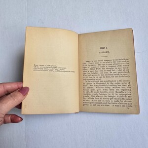 Peut inclure: Un livre ouvert avec des pages jaunies et vieillies. La page de gauche contient un po&egrave;me et la page de droite le titre "ESSAI L. HISTOIRE". Le texte est &agrave; l'encre noire. Le livre est tenu ouvert par une main.