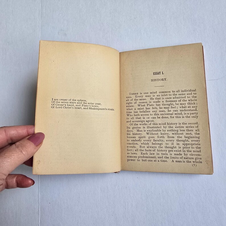 Peut inclure: Un livre ouvert avec des pages jaunies et vieillies. La page de gauche contient un po&egrave;me et la page de droite le titre "ESSAI L. HISTOIRE". Le texte est &agrave; l'encre noire. Le livre est tenu ouvert par une main.