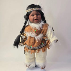 May include: A Native American doll with dark skin, black braided hair, and a headband. The doll wears a brown and white outfit with faux fur trim, beaded accents, and fringed details. The doll's expression is open-mouthed.