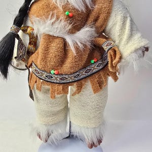 May include: A doll dressed in a Native American-inspired outfit. The doll features a brown top with white faux fur trim, cream-colored pants, and a long black braid. The outfit is adorned with colorful beads and patterned trim.