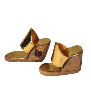 Puede incluir: Un par de sandalias de cuña de madera con detalles metálicos dorados. Las sandalias presentan una correa dorada texturizada y una base de madera. El tacón de cuña mide aproximadamente 7,6 cm de alto.