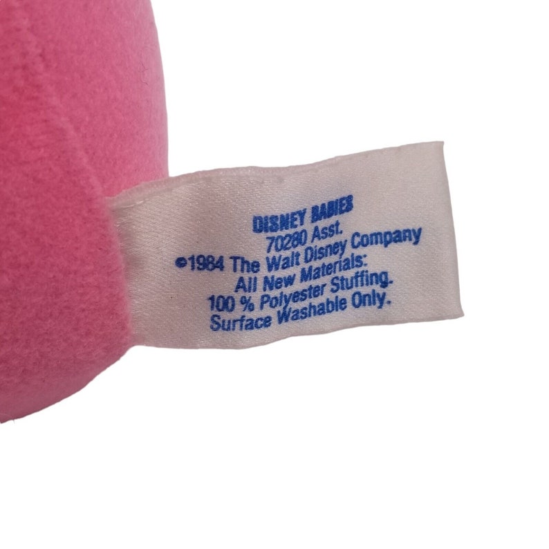 Puede incluir: Una etiqueta blanca con texto azul que dice "DISNEY BABIES 70280 Asst. &copy;1984 The Walt Disney Company All New Materials: 100% Polyester Stuffing. Surface Washable Only."