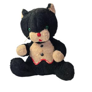 Puede incluir: Un peluche vintage de gato negro con ojos verdes, nariz roja y pecho y patas beiges. El gato tiene un ribete rojo alrededor del pecho y dos botones negros. El gato tiene un solo bigote.