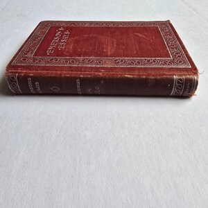 Peut inclure: Un livre ancien intitul&eacute; "Emerson's Essays" avec une couverture rouge et une bordure argent&eacute;e d&eacute;corative. La tranche du livre affiche le titre et les informations de la s&eacute;rie. Le livre est en bon &eacute;tat, montrant des signes d'&acirc;ge.