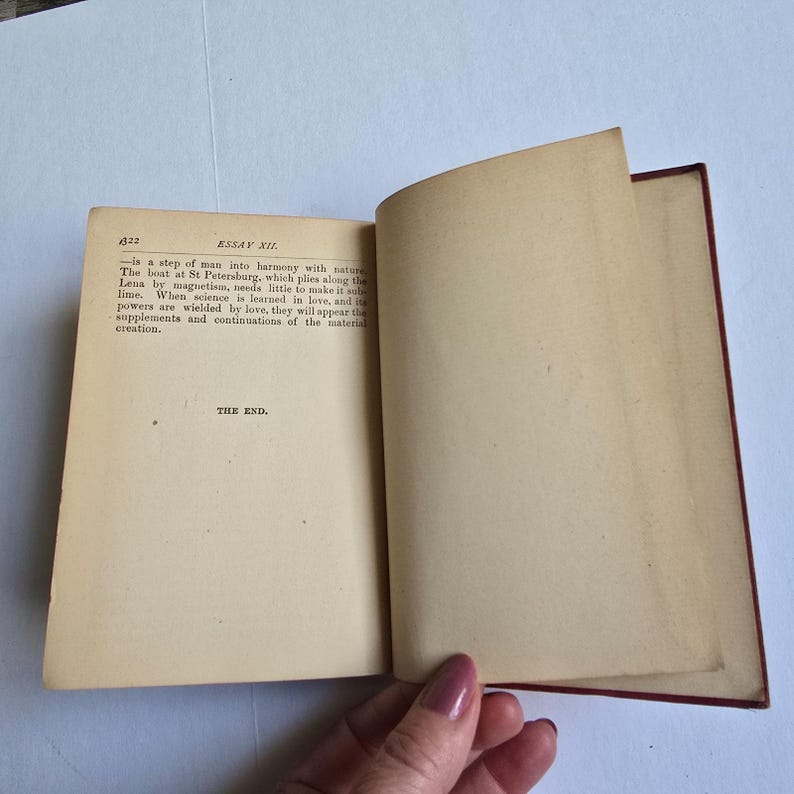 Peut inclure: Un livre ancien ouvert avec des pages jaunies. La page de gauche affiche du texte, dont "ESSAI XII" et "THE END." La couverture du livre est rouge fonc&eacute;, et une main est visible tenant le livre ouvert.
