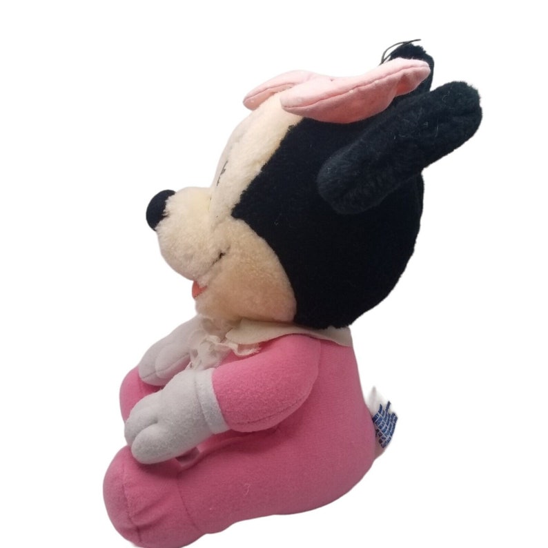 Puede incluir: Un juguete de peluche rosa y blanco de una Minnie Mouse beb&eacute; que lleva un vestido rosa y un lazo rosa. El juguete est&aacute; sentado con la cabeza girada hacia un lado.