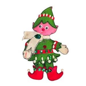 May include: A felt Christmas elf ornament. The elf has a green outfit with red accents, a white scarf, and red shoes. The elf's face is pink with black eyes and a red nose. The outfit is decorated with green sequins and white pearls.