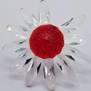 Könnte beinhalten: Eine klare Kristallblume mit einem leuchtend roten Zentrum. Die Blütenblätter sind facettiert und reflektieren das Licht. Die Blume ist ein dekorativer Gegenstand, möglicherweise eine Figur oder ein Ornament.
