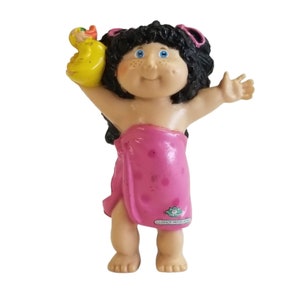 Puede incluir: Figura de Cabbage Patch Kids de una muñeca con cabello negro, ojos azules y pecas, envuelta en una toalla rosa con lunares. La muñeca sostiene un patito de goma amarillo. La figura está de pie con los brazos extendidos.
