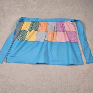 May include: A blue and white gingham apron with a patchwork design of green, yellow, orange, and pink squares. The apron has two long ties.