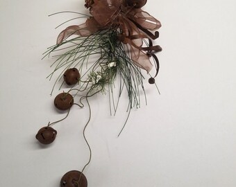 May include: A brown and green floral arrangement with small, rusty bells hanging from a wire. The arrangement includes pine needles, brown ribbon, and small white flowers.