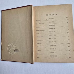 Peut inclure: Livre ouvert avec une tranche rouge, affichant la table des mati&egrave;res. La page r&eacute;pertorie des essais sur des sujets tels que l'histoire, la confiance en soi et l'amour. Le livre a un aspect fan&eacute; et vieilli, avec un tampon sur la page de gauche. Le texte est en noir.