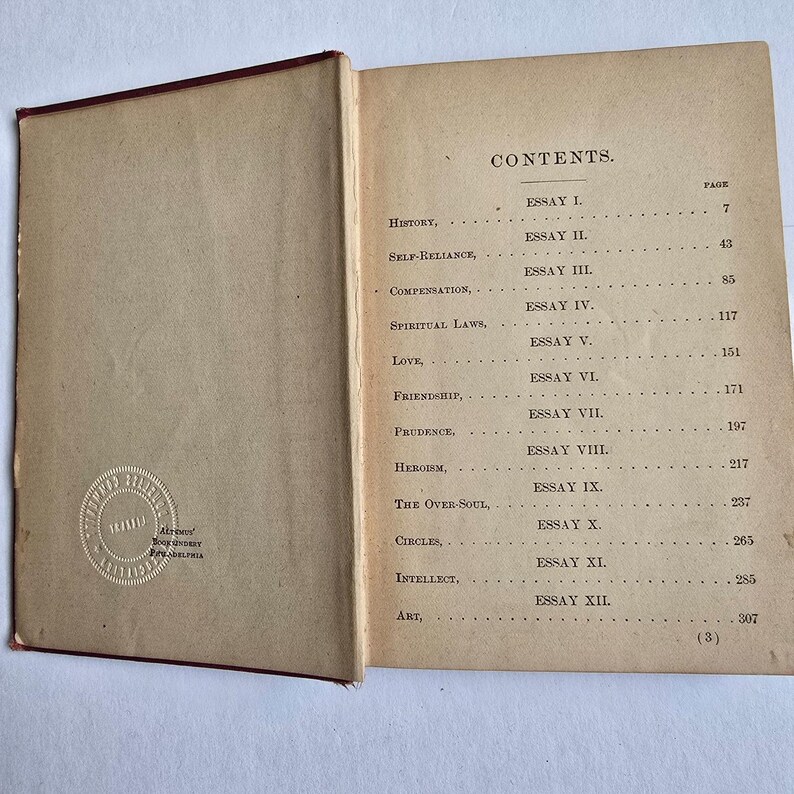 Peut inclure: Livre ouvert avec une tranche rouge, affichant la table des mati&egrave;res. La page r&eacute;pertorie des essais sur des sujets tels que l'histoire, la confiance en soi et l'amour. Le livre a un aspect fan&eacute; et vieilli, avec un tampon sur la page de gauche. Le texte est en noir.