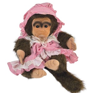 Puede incluir: Una muñeca de peluche de chimpancé con un vestido y un gorro de cuadros rosas. La muñeca tiene pelo marrón, un chupete y está sentada. El vestido tiene ribetes y volantes blancos. La cara de la muñeca es de color marrón claro con ojos oscuros.