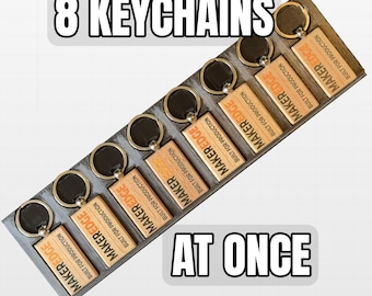 May include: Eight rectangular wooden keychains arranged in a diagonal row, each featuring a metal ring and the black printed text MAKER EDGE BUILT FOR PRODUCTION. The text 8 KEYCHAINS AT ONCE is overlaid on the image in bold white letters.