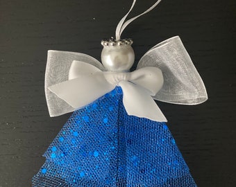Puede incluir: Un adorno de ángel azul y blanco con un lazo blanco y una corona de cuentas plateadas. El ángel está hecho de tul y tiene un acabado brillante.